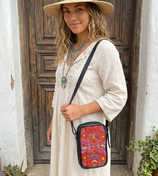 Woman wearing a wide-brimmed hat with feather accents and a patterned bag, standing in front of a wooden door.