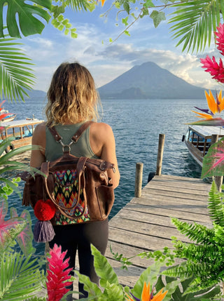 Person with a colorful backpack standing on a dock by a lake with a mountain in the background, surrounded by tropical plants and flowers.