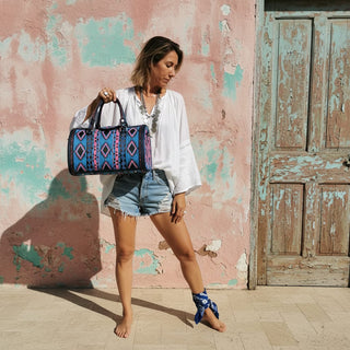 Woman holding a patterned bag in front of a textured pink wall and wooden door.