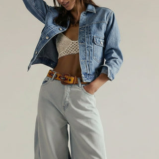 Woman wearing a denim jacket, white top, and light gray pants against a plain background