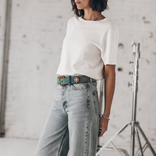 Woman wearing a white top and light blue jeans standing in a studio with a white brick wall background.